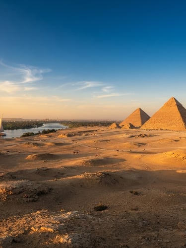 Wide panoramic view of the Giza Pyramids at golden hour, uncrowded, warm low-angle sunlight casting long shadows, a single luxury dahabiya visible on the distant Nile, cinematic, 35mm lens.