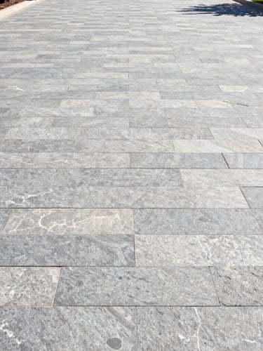 Low-angle wide shot of a newly completed interlocking stone driveway in bright morning sunlight, clean slate gray and limestone tones, sharp architectural lines, high-contrast details.