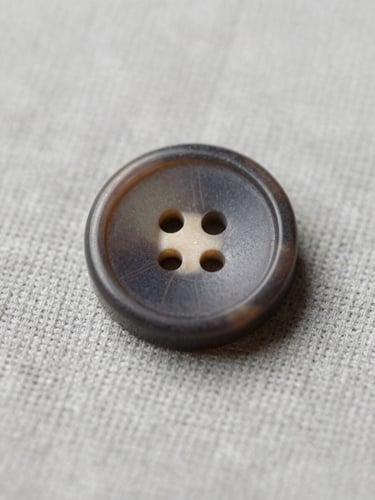 Macro photograph of a single exquisite dark horn button on a light gray linen fabric, showing its unique natural grain and precise 20mm caliber, soft diffused studio lighting.