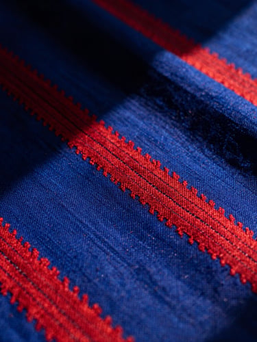 Macro shot of hand-loomed silk Ikat fabric with vivid lapis blue and madder red patterns, dramatic natural sunlight casting deep shadows, showcasing the rich tactile texture of the weave.