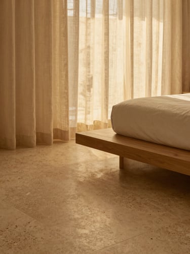 Architectural detail of a minimalist bedroom suite, warm travertine floor, a single low oak platform bed, soft golden-hour light filtering through sheer linen drapes.