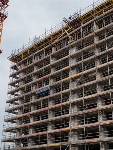Wide construction site at daylight, complete scaffolding system spanning a multi-story building facade to the right, steel frames under load, workers visible at mid-level, overcast sky revealing joint and brace detail