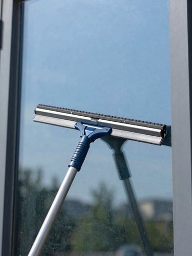 Detail shot of a professional squeegee wiping a window clean, leaving a streak-free reflection of blue sky and green trees, bright daylight, 35mm lens