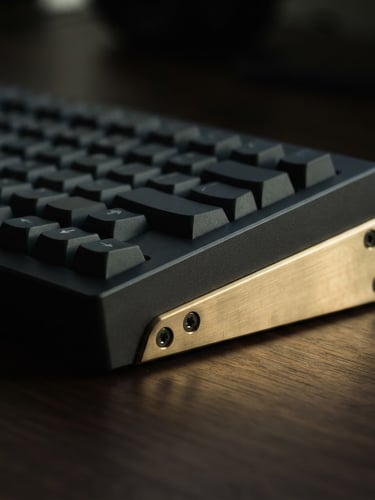 Dramatic side profile of a heavy charcoal aluminum keyboard chassis with a solid brass weight, resting on a dark desk, low-key directional window light casting rich shadows, macro lens, shallow depth of field