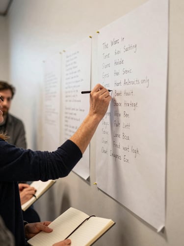 Wide shot of a facilitator's hand writing a key phrase on a large paper sheet pinned to a wall, other participants' hands and notebooks visible at the frame edge, warm diffused indoor light, genuine in-session moment captured from a low side angle