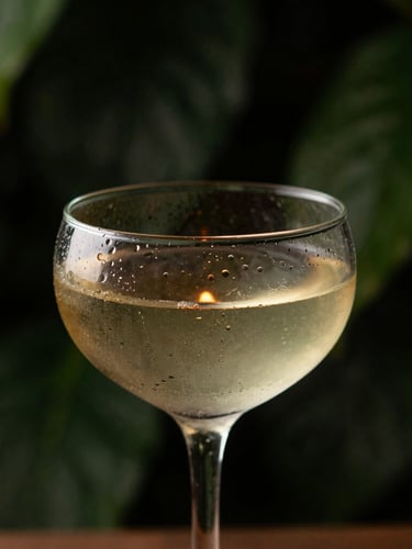 Close-up of a single cocktail in delicate glassware, condensation on glass, warm candlelit glow, deep shadow-heavy forest green background, shallow depth of field.