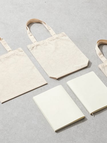 Minimalist studio flat-lay of custom organic cotton tote bags and notebooks arranged on a light gray concrete surface, bright high-key lighting, soft shadows