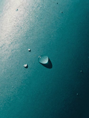 Tight abstract macro crop of a matte-finished deep teal resin surface with water droplets glistening under harsh direct sunlight, sharp high-contrast shadows.