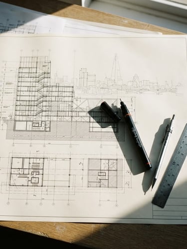 Top-down flat lay of a clean drafting table in London, stark morning side-light casting sharp shadows across architectural blueprints and a technical pen.
