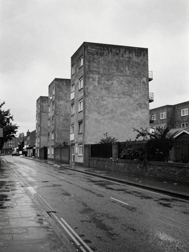 B&W wide shot of wet asphalt streets in Canning Town under overcast, bleak daylight, brutalist concrete housing estates looming in the background.