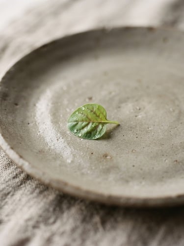 Extreme close-up of a minimalist ceramic plate with a single micro-green leaf, soft natural side light, raw linen napkin texture, muted warm gray tones, shallow depth of field, 35mm film aesthetic