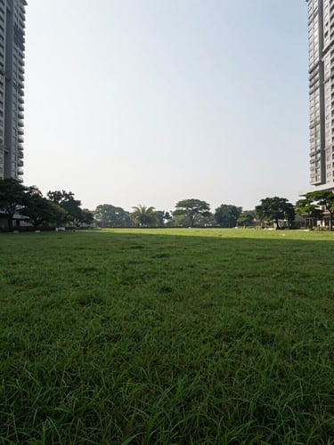 Low-angle panoramic view of the open ground plane at Godrej South Estate, no vehicles, deep green planted landscape stretching across the foreground, the base of two towers visible at the frame edges, clear morning overcast light flattening shadows to show the scale of empty ground
