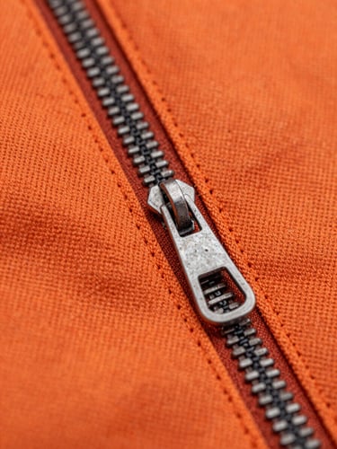 Close-up macro shot of double-stitched heavy-duty orange canvas fabric, focus on a rugged steel zipper and reinforced seam, industrial lighting.