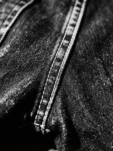 Close-up of distressed black denim fabric with visible white stitching, harsh direct sunlight, deep shadows, high-contrast monochrome, 35mm film grain, side view, wide angle.