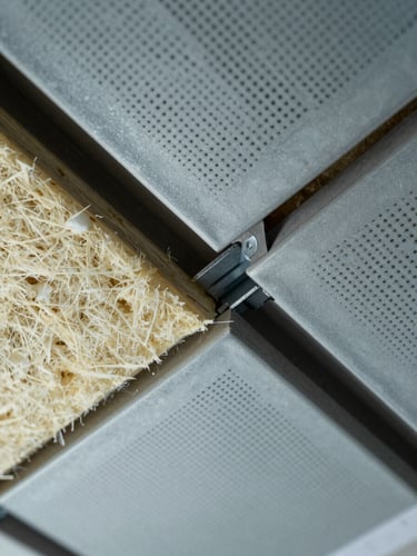 Extreme close-up overhead view of a wall-to-ceiling acoustic panel joint showing mineral wool substrate, perforated metal face, and structural clip detail, cool diffused daylight from above, no people, construction drawing partially visible at edge