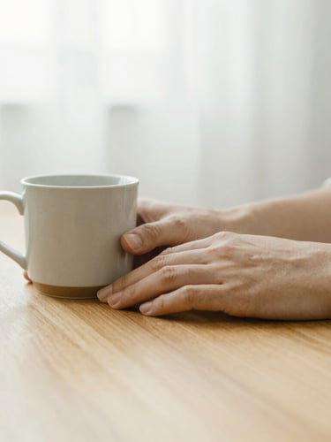 A close-up of hands resting on a warm wooden table next to a ceramic mug, soft overcast daylight, shallow depth of field, calm and quiet atmosphere.