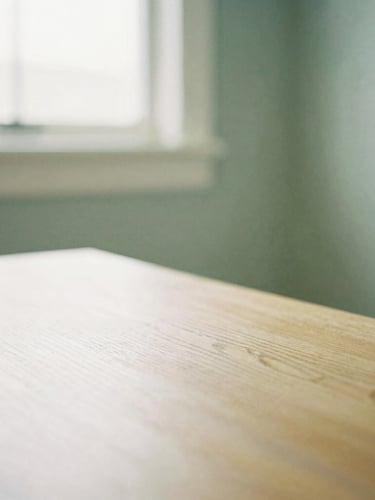 A soft-focus close-up of a warm cream consultation room, natural light streaming through a window onto a wooden table, sage green accent wall, shallow depth of field, 35mm