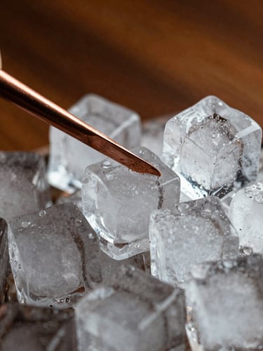 Macro shot of large clear ice cubes being carved with a copper tool, dramatic side lighting, rich warm tones of wood and copper