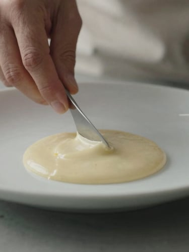 Close-up of blurred hands plating a minimalist dish in a quiet Scandinavian-style kitchen, focus on the texture of the sauce, soft side lighting.