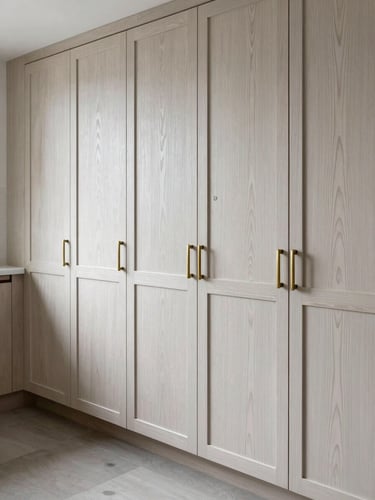 Close environmental shot of a completed kitchen — inset cabinet doors in natural white oak, grain running vertically, hardware in aged brass, photographed in north-facing daylight. Empty room, no staging. Wide portrait framing showing floor-to-ceiling cabinetry wall, emphasis on the flush reveal lines and consistent grain pattern across panels.