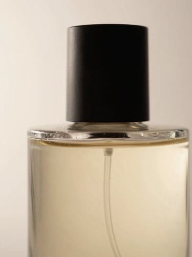 Extreme close-up of a cylindrical perfume bottle, raking light from the left revealing the curve of the glass and the texture of the cap, matte surface, no warmth, pale background
