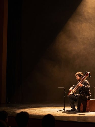 Wide archival concert photograph, a classical sitar performer seated on stage in a dimly lit mehfil hall, warm amber stage light falling from the upper right, the musician's profile visible against deep shadow, audience silhouettes in the near foreground, generous dark negative space to the left third of the frame
