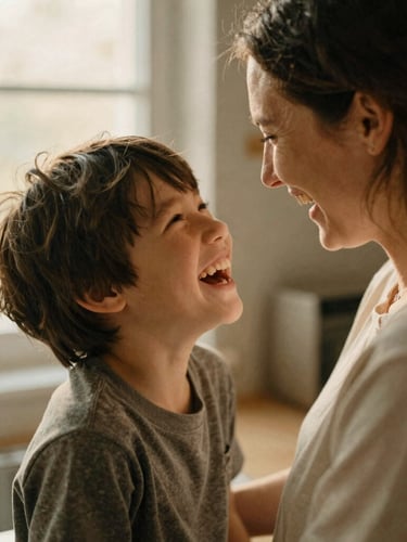 Candid medium close-up of a mother and her neurodivergent son laughing together in a warm, sunlit kitchen, soft natural window light, muted earth tones, 35mm film grain