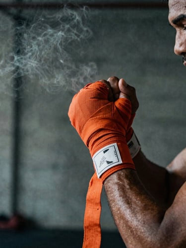 Dramatic side-lit close-up of a kickboxer wrapping their hands with orange handwraps in a raw, dark concrete gym, atmospheric smoke, 35mm photography
