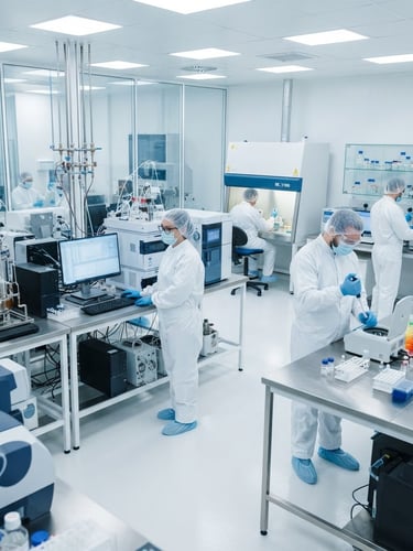 Wide shot of a sterile pharmaceutical laboratory with scientists in cleanroom suits operating high-precision analytical equipment, bright clinical lighting, blue and white tones.