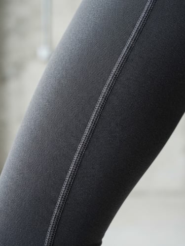 Bright studio macro shot of model wearing charcoal grey technical leggings, focusing on the flatlock stitching detail and dense 4-way stretch fabric texture, crisp high-contrast lighting.