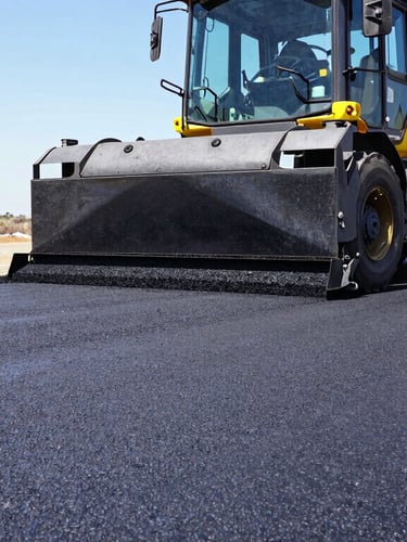 Low-angle heroic shot of a heavy asphalt paving machine laying textured black hot-mix asphalt under bright South African daylight, safety yellow accents visible.
