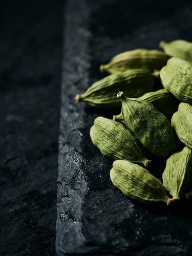 Macro close-up of vibrant green cardamom pods, dramatically side-lit, rich deep tones, 35mm, elegant luxury aesthetic, on a dark stone surface, with generous negative space to the left.