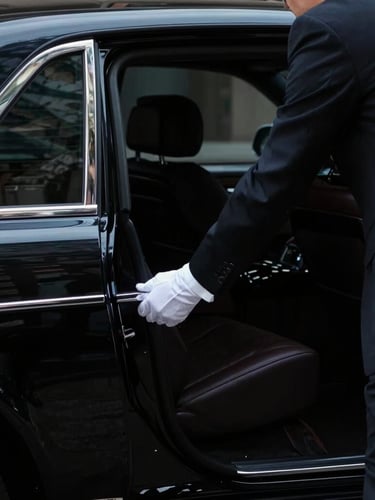 Portrait image of a chauffeur in a sharp dark suit, with white gloves, opening the rear door of a black executive sedan, interior cabin visible, low-key dramatic lighting, sharp focus on the hands and door.