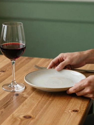 Warm editorial photography of a rustic wooden restaurant table with soft natural side-light, a glass of red wine, and hands arranging a ceramic plate, 35mm