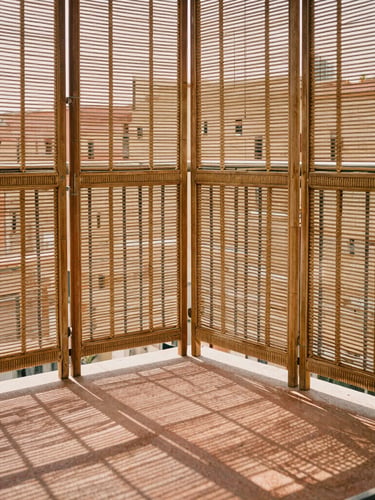 A sunny urban balcony with a structural rattan screen blocking the wind, casting intricate geometric shadows over a warm terracotta floor.