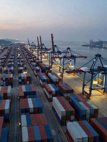 Panoramic overhead aerial shot of a major international container port at dusk, rows of stacked cargo containers stretching to the left horizon, electric-blue crane lights active on the right third, cool industrial haze, vast scale of global freight infrastructure