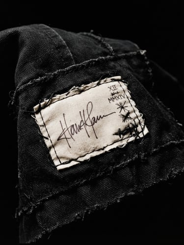 Stark editorial fashion photography of a hand-signed artist label stitched onto thick canvas-weight black textile, harsh dramatic studio lighting, deep shadows