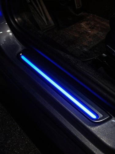 Moody night photography of a sports car door sill glowing with vibrant electric indigo LED light, reflecting off dark metallic door frame.