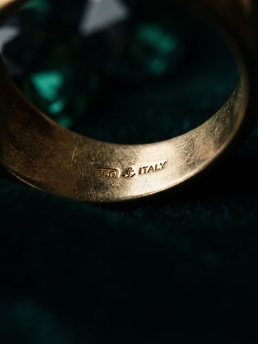 Macro editorial photograph of hallmarked gold edges catching dramatic low-key spotlight, rich emerald shadows, shallow depth of field, 85mm lens