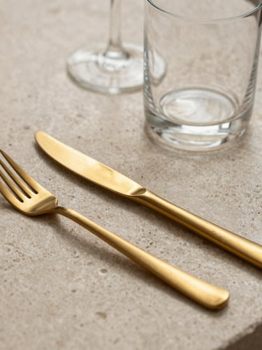 Macro detail of a bespoke textured stone table setting, sharp directional spotlighting highlighting gold-brushed cutlery and minimalist glassware.