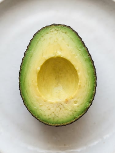 Top-down macro shot of sliced avocado showing rich green-to-yellow gradient flesh on a clean off-white surface, soft morning light