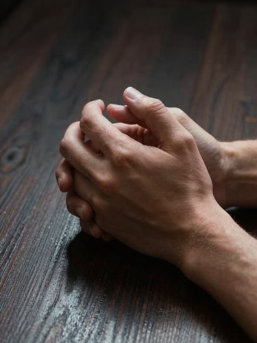 High-contrast close-up of a person's hands clasped in quiet reflection over a dark, textured wood surface. Deep side-lighting, heavy shadows, no screens or technology visible.