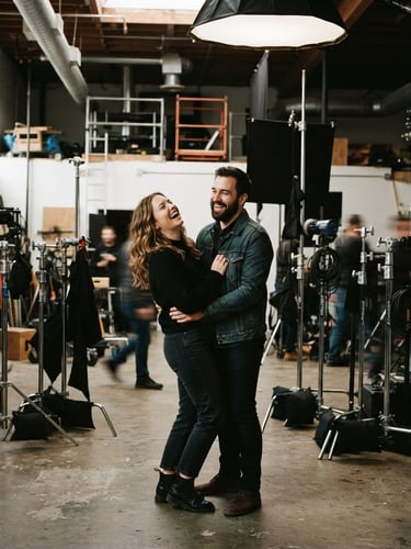 Cinematic wide shot of a couple laughing candidly in an industrial studio space, soft shadows, warm film grain, visible C-stands in the background, 35mm photography