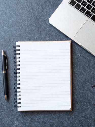 Overhead flat-lay of an organized desk with a notebook, pen, and laptop, bright natural daylight, sharp focus, 35mm