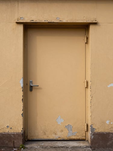 Wide environmental shot of a weathered industrial doorway, peeling ochre paint layers, diffused overcast daylight casting soft even shadows, architectural texture dominant, no human subjects, close compositional detail showing surface decay and geometry