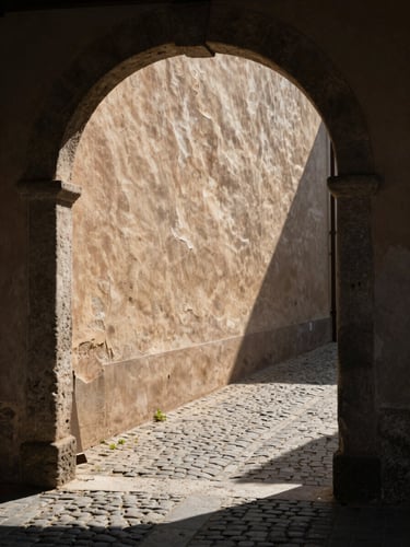 Portrait-format frame looking through a narrow stone archway into a sun-drenched cobblestone lane, deep shadow on the near wall, warm raking light catching the far wall's plaster texture, no people, strong geometric framing, natural morning light
