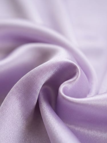 Macro close-up of high-sheen lavender satin fabric draped elegantly with soft natural daylight casting gentle shadows, bright editorial photography, shallow depth of field