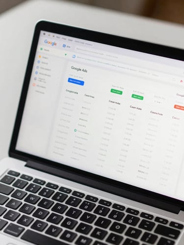 Close-up overhead view of a laptop screen displaying a live Google Ads campaign dashboard with click-through rate and cost-per-click columns visible, on a clean desk under even north-facing daylight, keyboard partially in frame