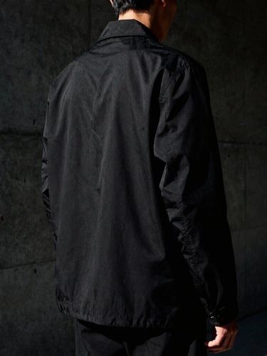 Low-key, high-contrast model shot from behind, showing the architectural drape of a structural black jacket against a dark concrete wall, harsh side-lighting, 35mm film