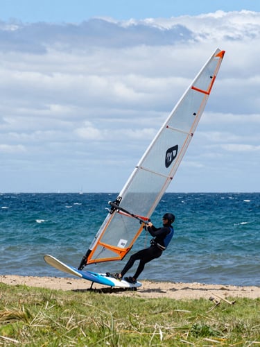 A close-up action shot of a windsurfer rigging a sail on the grassy bank of Christchurch Harbour, overcast maritime sky, rich ocean blues.